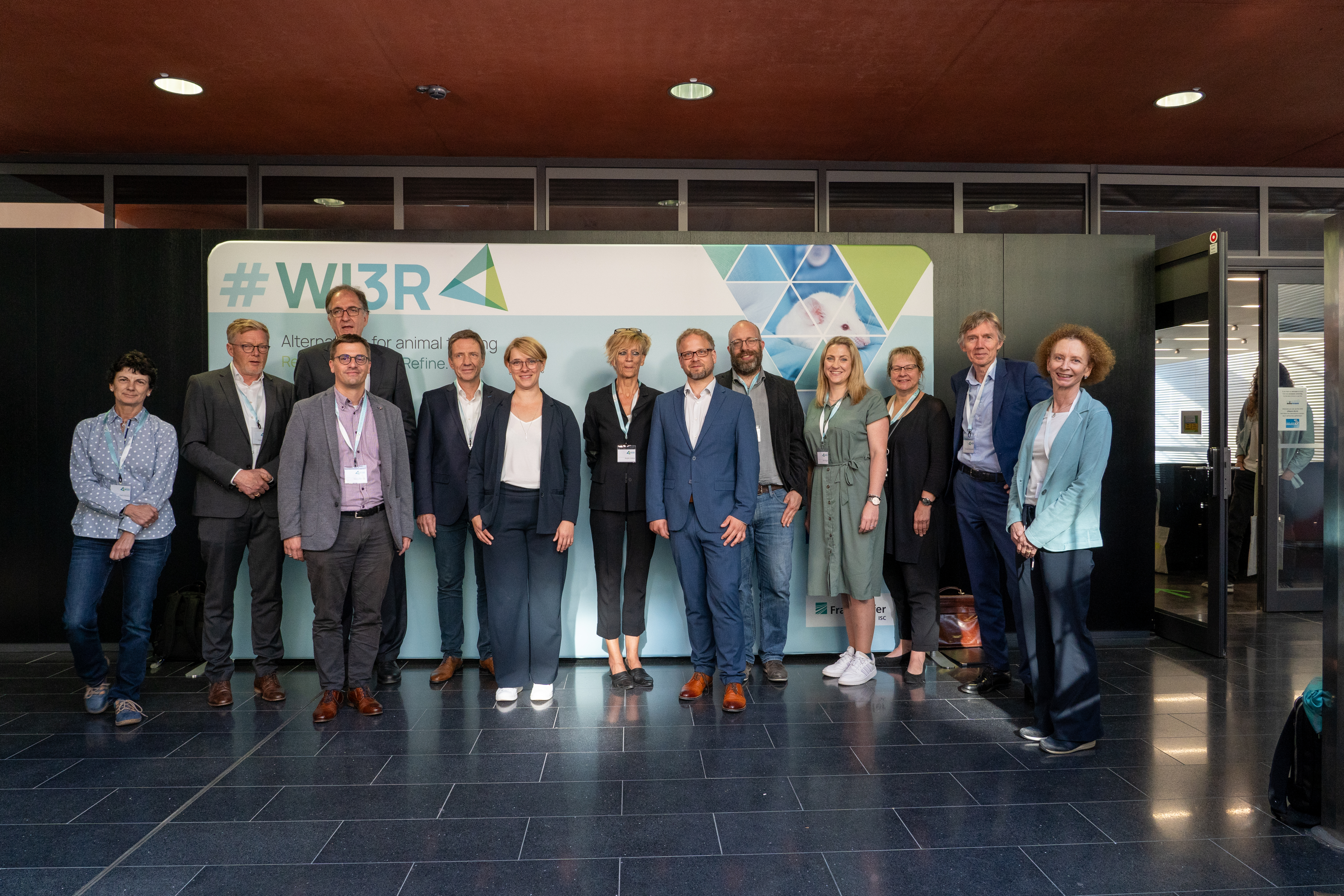 Gruppenbild mit allen an Preisverleihung und Rückblick beteiligten Personen. 13 Menschen stehen vor einer Fotowand des WI3R-Syposiums, 