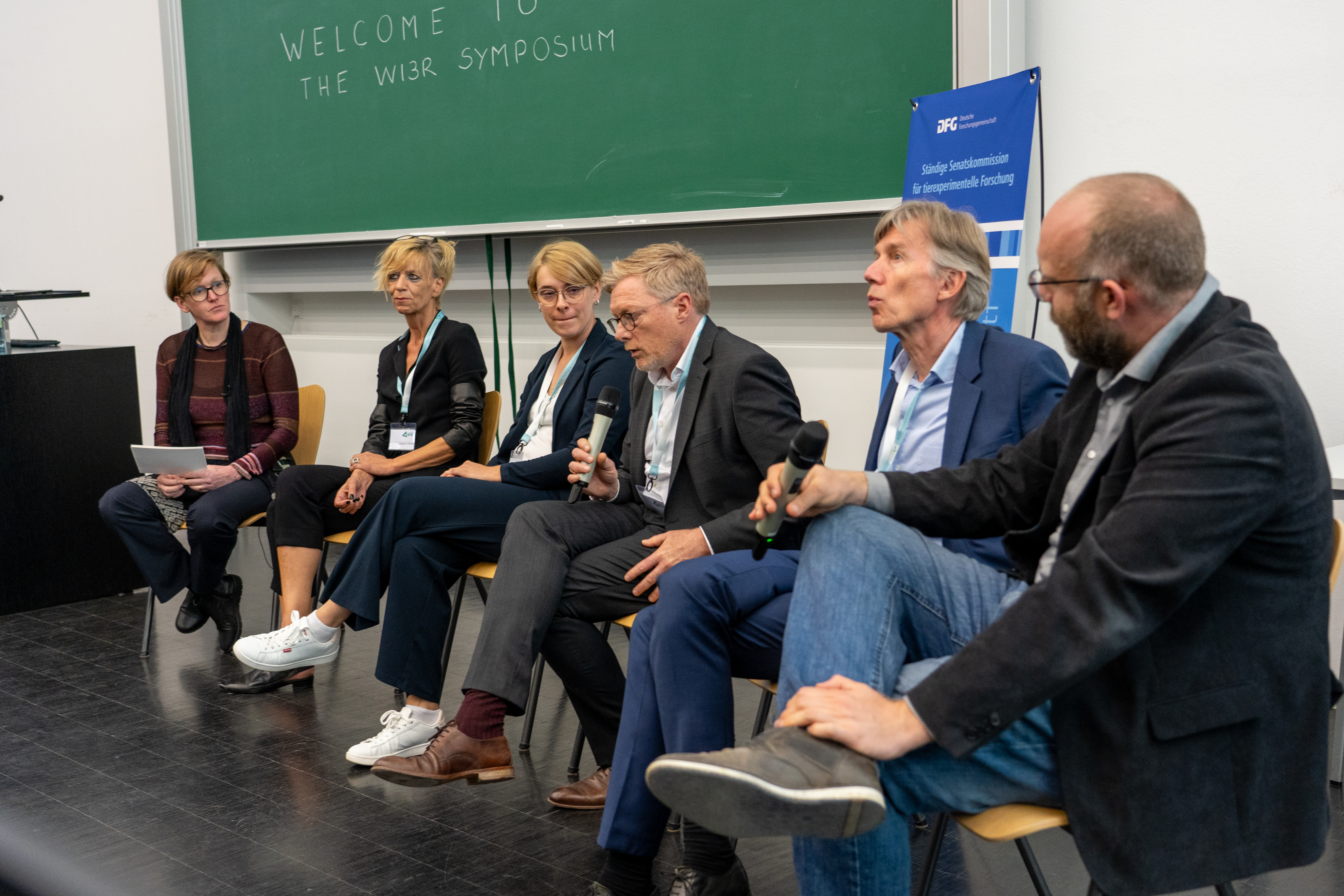 Sechs Personen diskutieren bei der Podiumsdiskussion miteinander. Sie sitzen in einem Hörsaal vor einer Tafel.