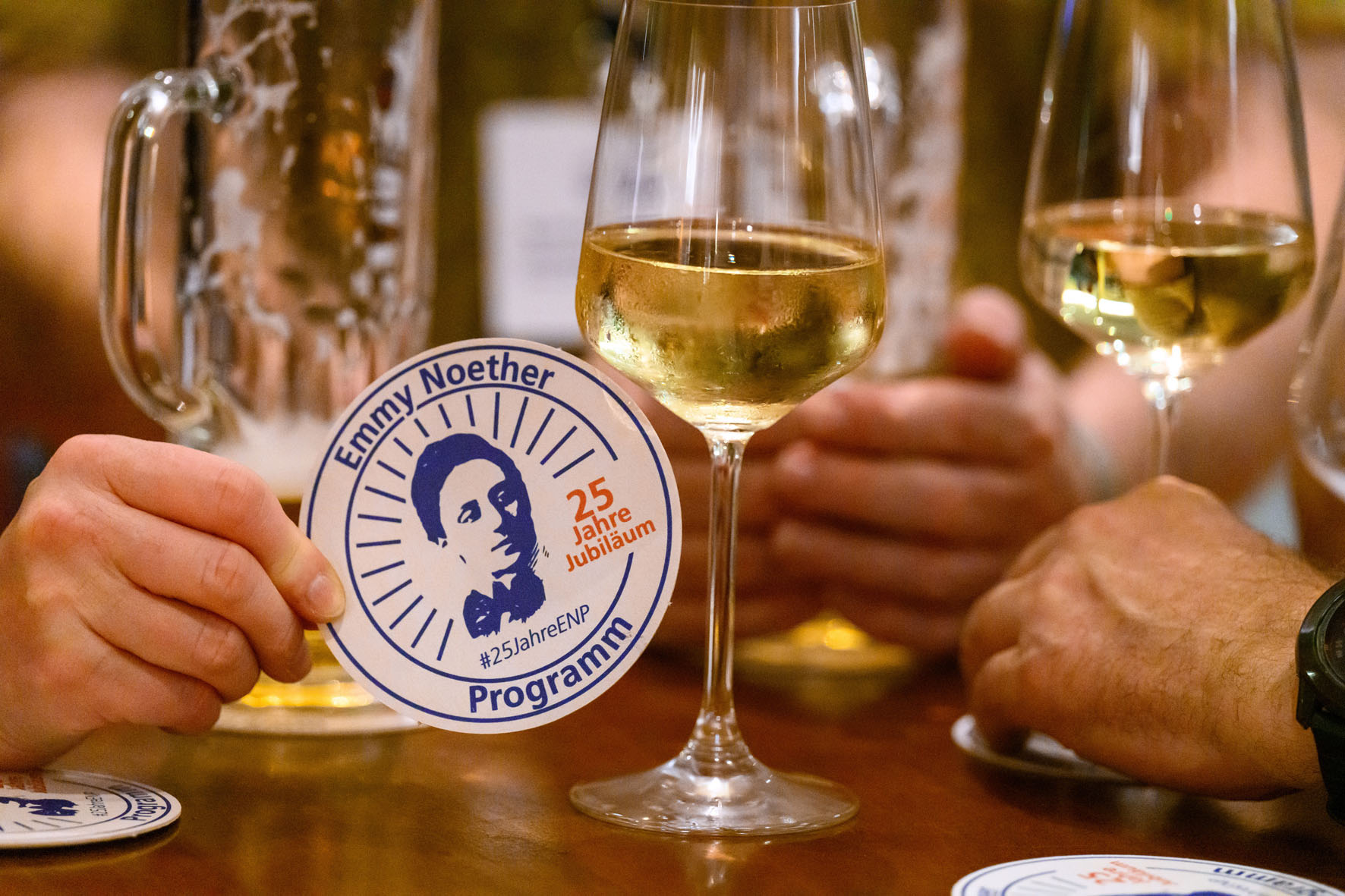Ein Bierdeckel bedruckt mit "25 Jahre Emmy Noether-Programm" vor einem Weinglas