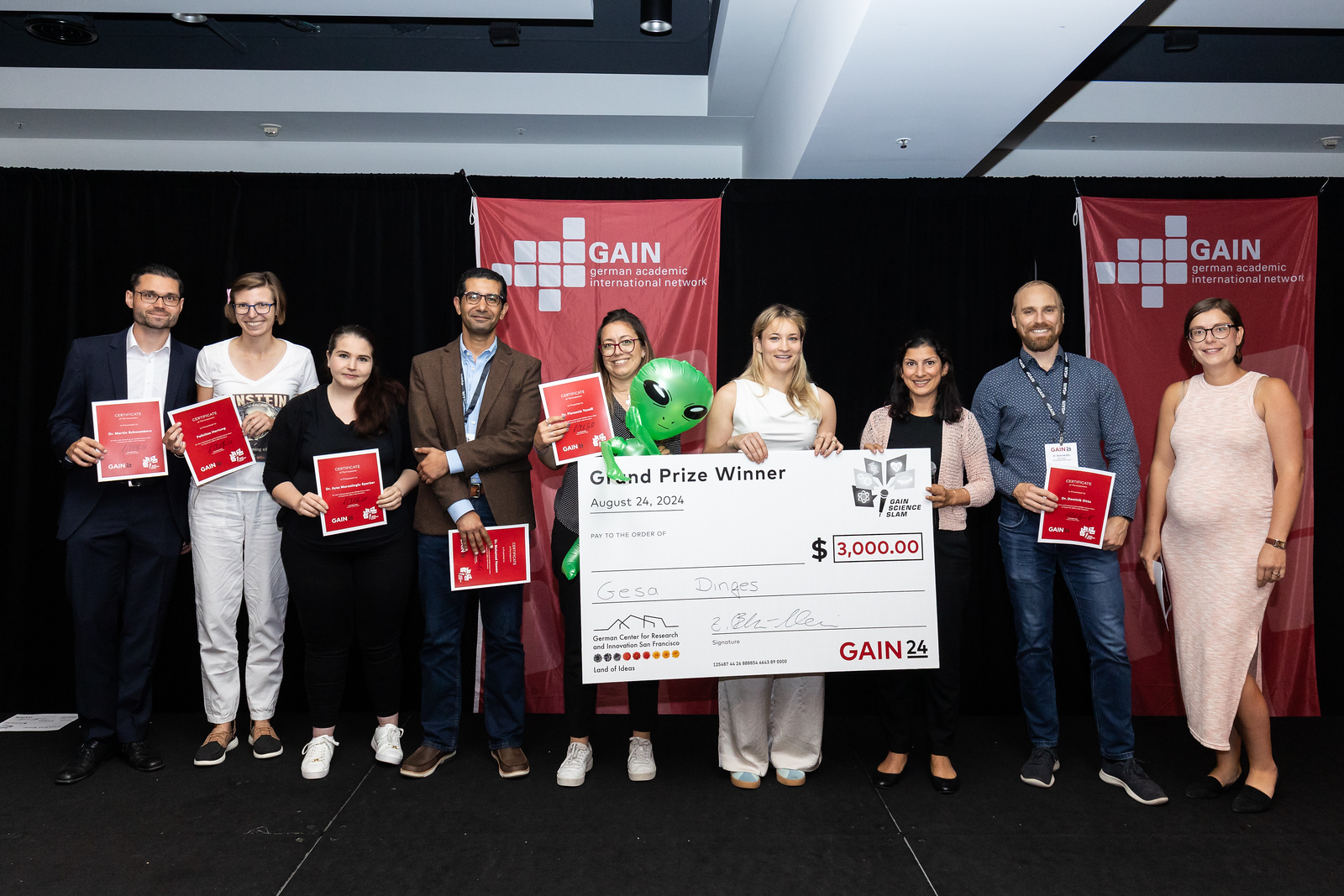 The winner holds a large check in front of her, the other participants stand next to her