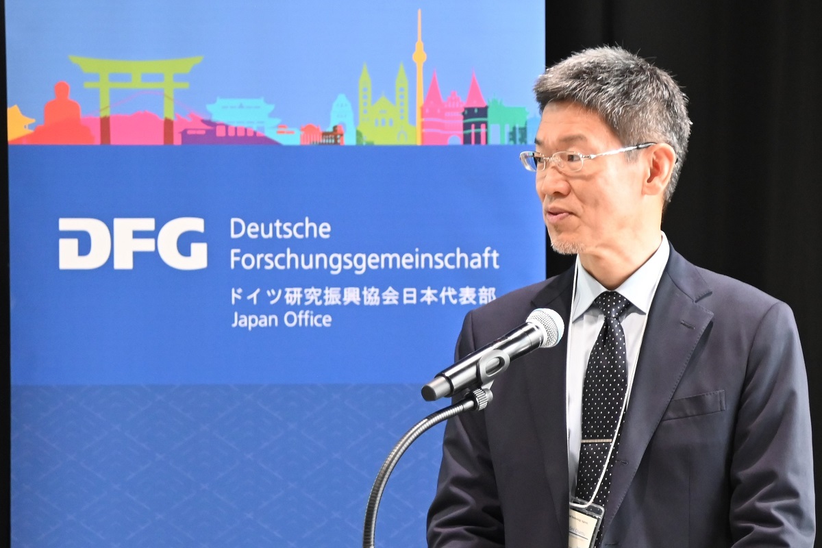 Osamu Kobayashi speaks to the guests from the podium. A DFG stand-up display can be seen in the background.