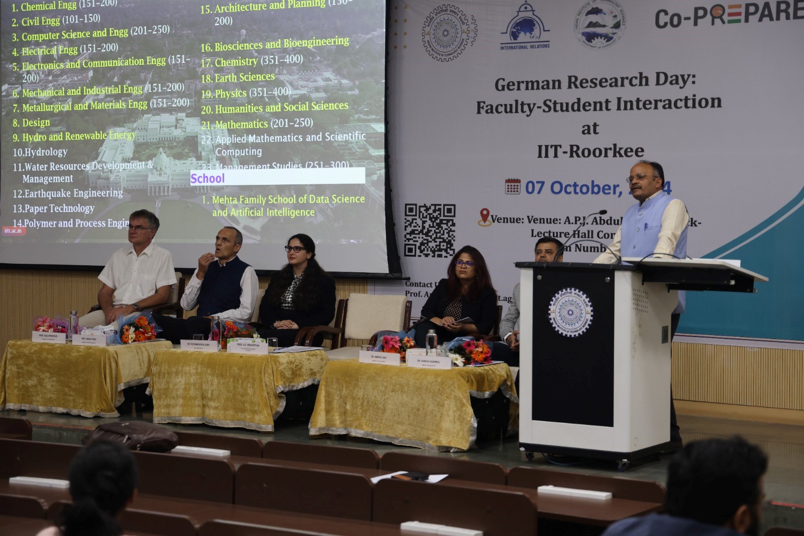 The picture shows the podium with several lecturers in front of a presentation which reads "German Research Day".